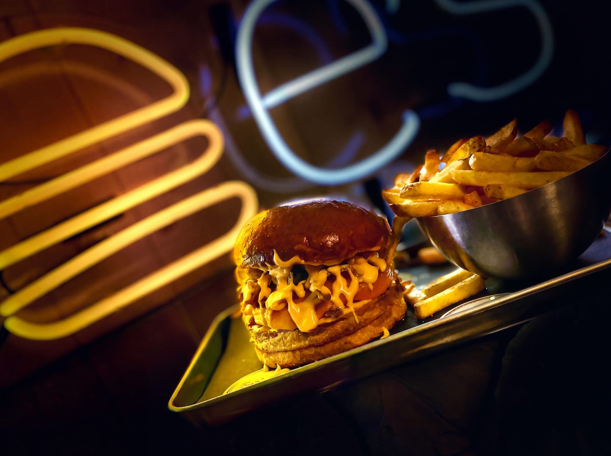 Burger mit Cheddar und Pommes auf einem Tablett vor gelbem Neonlicht